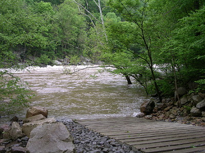 [This image taken from the top of the boat ramp which is a series of planks parallel to the water atop a bed of crushed rock. The water level covers the bottom of the ramp and whitewater is visible at the river's opposite edge.]